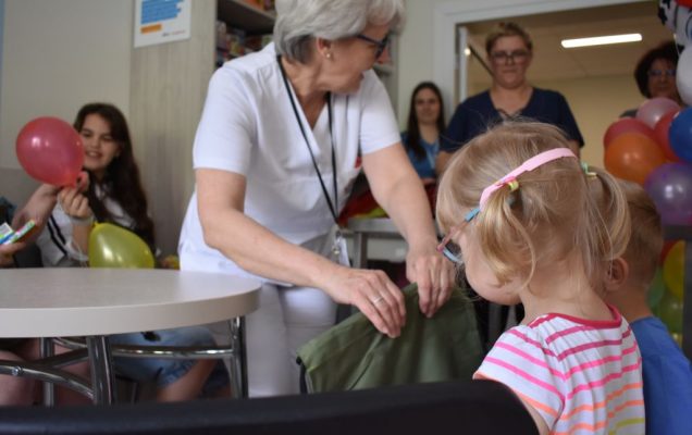 Pracownica szpitala i małą dziewczynka. Kobieta siedzi na krzesełku i wręcza prezenty.