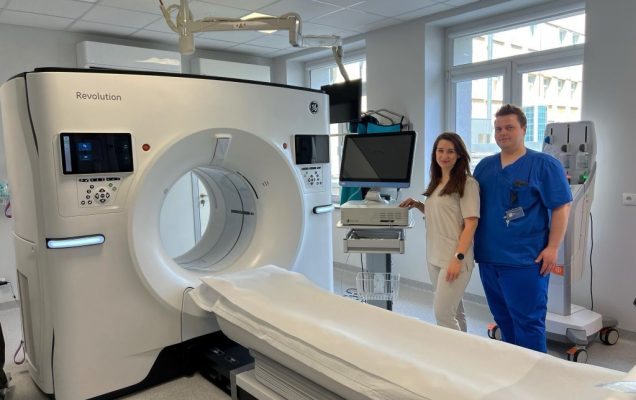 Dwóch pracowników służby zdrowia stoi obok tomografu komputerowego w sali szpitalnej wyposażonej w monitory i sprzęt medyczny.