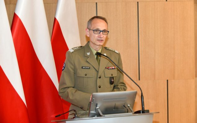 Dyrektor WIM-PIB Grzegorz Gielerak na auli WIM przy mównicy, za nim flagi Polski.
