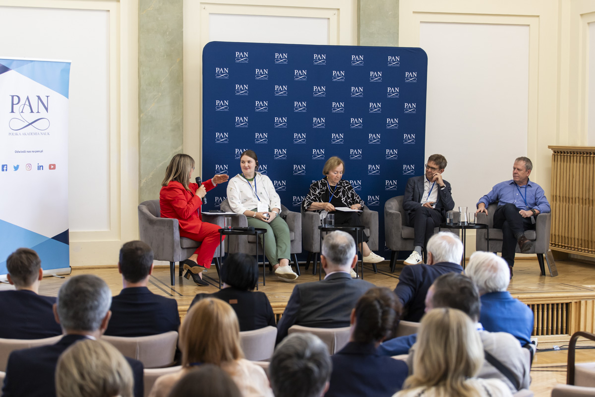 Panel dyskusyjny konferencji, siedzący prelegenci z animi tablica z logotypami PAN.
