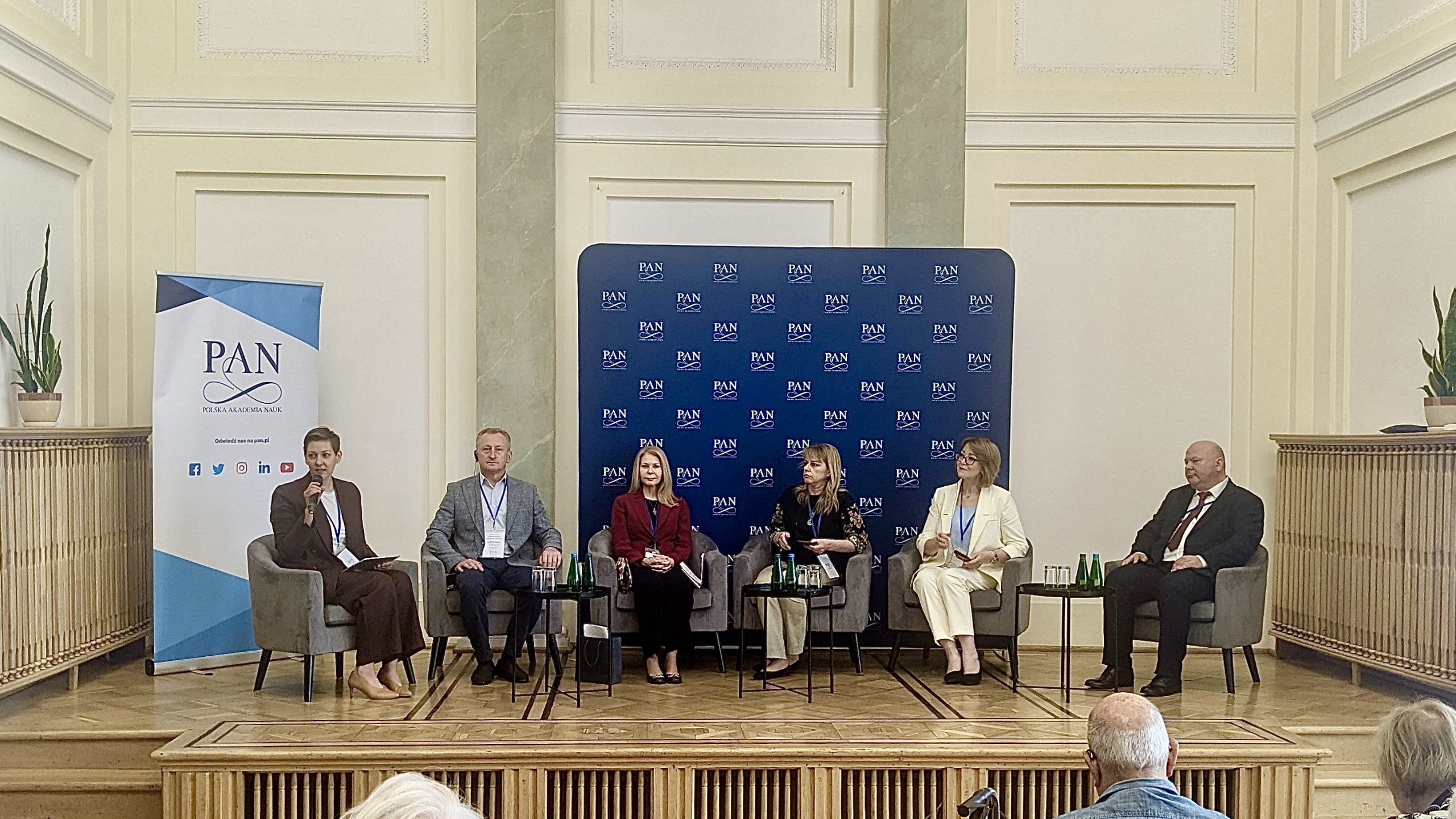 Panel dyskusyjny konferencji, siedzący prelegenci z animi tablica z logotypami PAN.