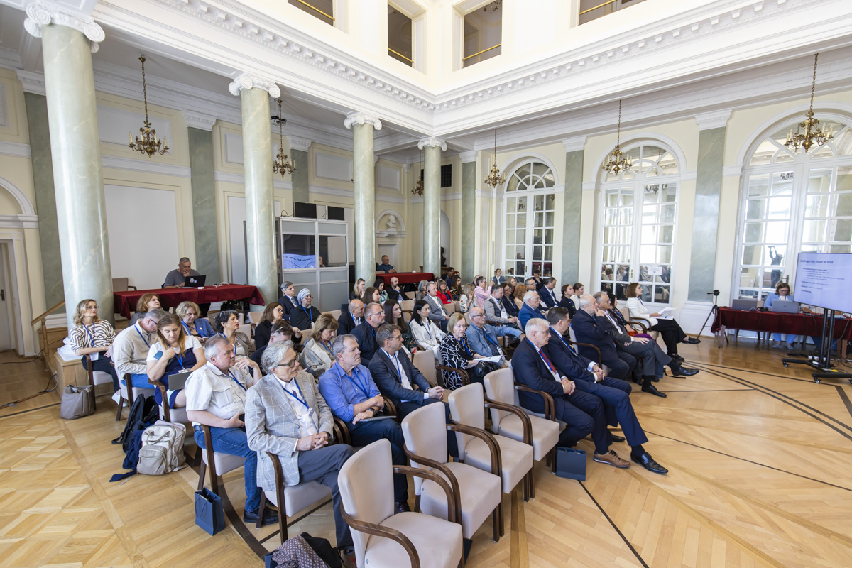 Uczestnicy konferencji siedzący w zabytkowej sali z kolumnami.