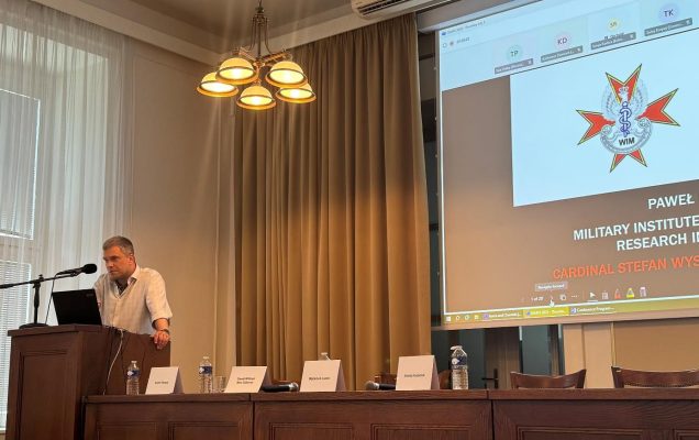 Prelegent podczas wykładu na konferencji. Za jego plecami wyświetlona prezentacja a na niej logo WIM.