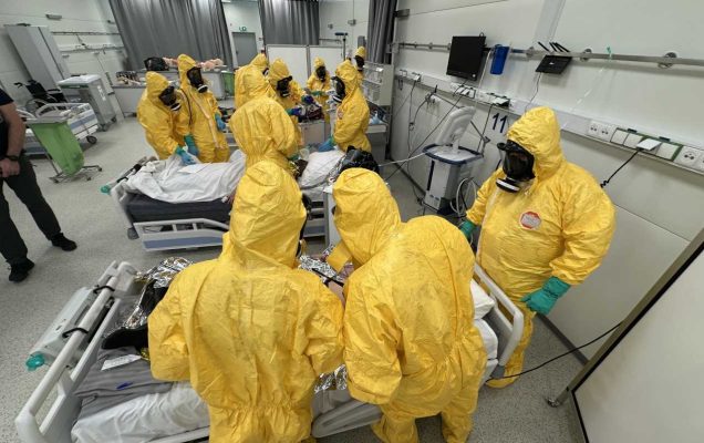 Medycy w żółtych kombinezonach i maskach przeciwgazowych podczas kursy CBRN w żółtych kombinezonach na sali szpitalnej.