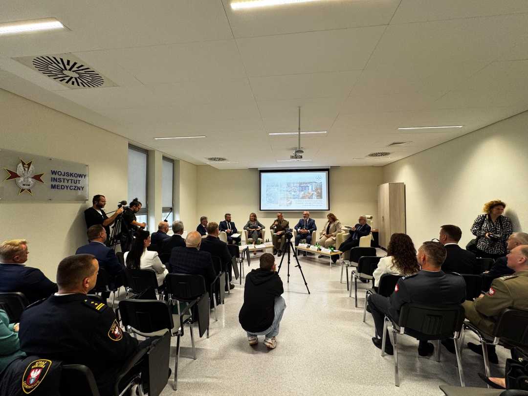 Sala konferencyjna pomiędzy rzędami krzeseł, po środku kucający dziennikarz robi zdjęcie uczestnikom debaty.