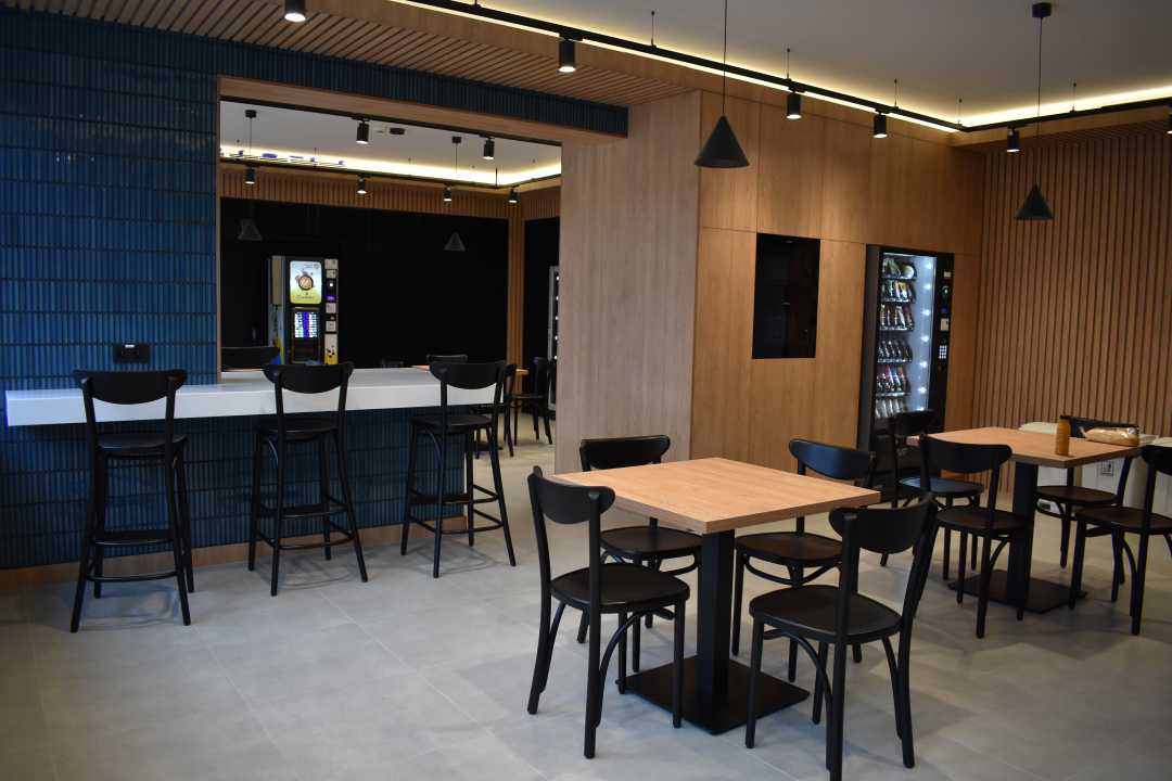 Na pierwszym planie dwa kwadratowe małe stoliki z krzesełkami. Za nimi długi stół z krzesłami barowymi. Pomieszczenie ma beżową posadzkę, drewniane ściany oraz głęboko niebieską glazurę na części ścian.
