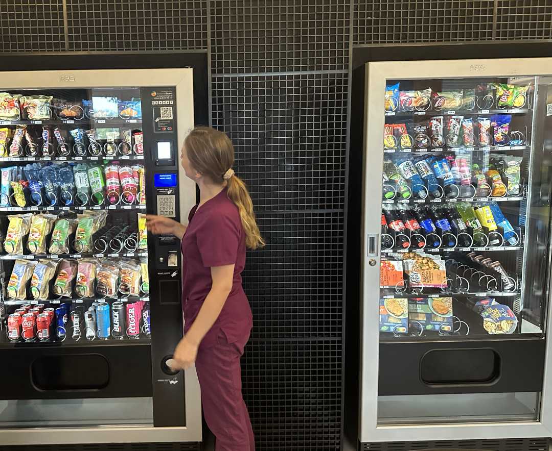 Dwa automaty do sprzedaży przekąsek. Przy jednym z nich stoi młoda pracowniczka WIM w bordowym uniformie i zapoznaje się z zawartością automatu.