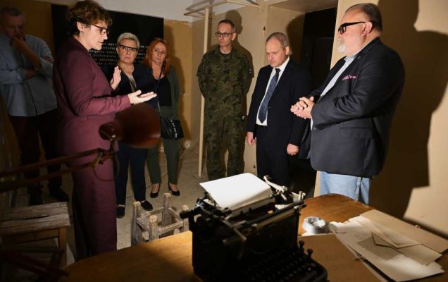 Pracownicy WIM w sali Muzeum Żołnierzy Wyklętych. Małą sala muzealna odtwarzającą pomieszczenie w warunkach wojennych. Na starym biurku rozłożone dokumenty i stara maszyna do pisania.