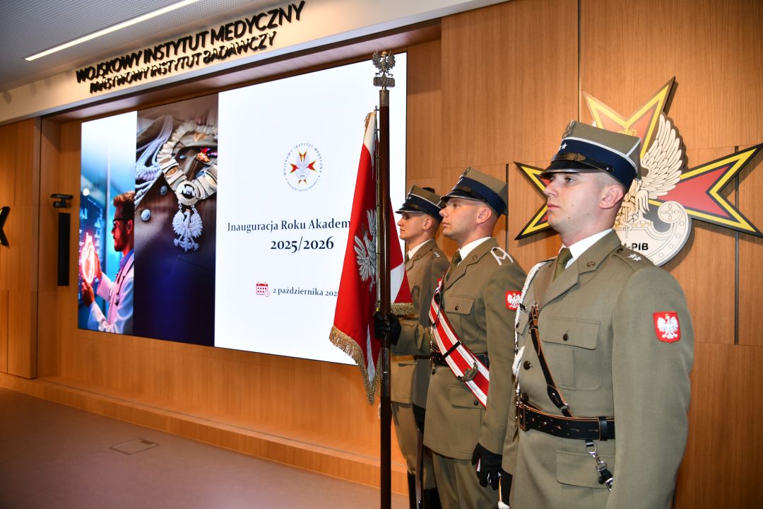 Inauguracja Roku Akademickiego w WIm-PIB. Aula WIM-PIB, trzech żołnierzy w mundurach galowych stoi na baczność z sztandarem Polski, obok nich wyświetlony napis Inauguracja Roku Akademickiego 2025/2026.