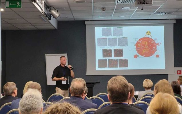 Sala wykładowa, ujęcie z tyłu sali uczestników siedzących na sali konferencyjnej podczas wykładu. Przed siedzącymi stojący prelegent przy prezentacji wyświetlanej z rzutnika.