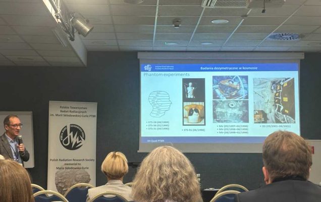 Sala wykładowa, ujęcie z tyłu sali uczestników siedzących na sali konferencyjnej podczas wykładu. Przed siedzącymi stojący prelegent przy prezentacji wyświetlanej z rzutnika.