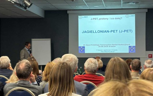 Sala wykładowa, ujęcie z tyłu sali uczestników siedzących na sali konferencyjnej podczas wykładu. Przed siedzącymi stojący prelegent przy prezentacji wyświetlanej z rzutnika.
