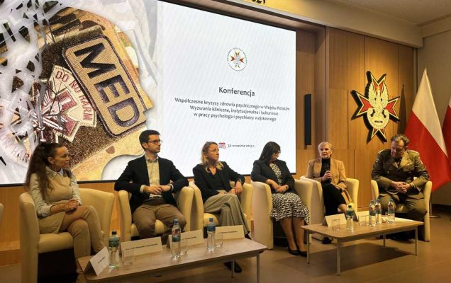 Prelegenci siedzący w fotelach podczas panelu dyskusyjnego. Za ich plecami na dużym wyświetlaczu strona tytułowa konferencji. Po prawej stronie stojak z flagą RP a nad nim Logo WIM-PIB.