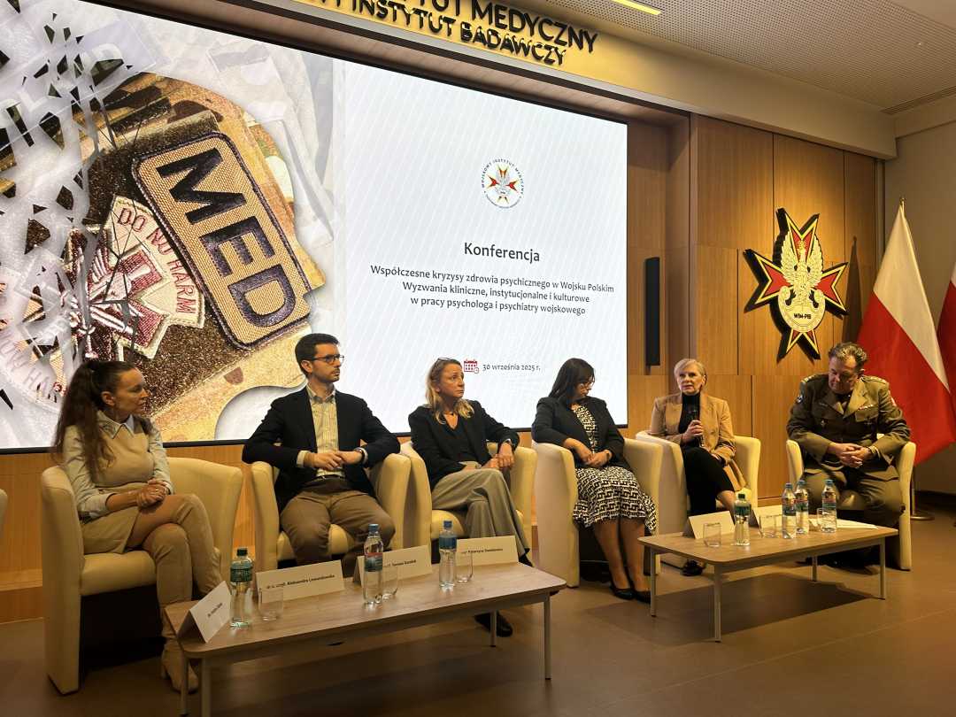 Prelegenci siedzący w fotelach podczas panelu dyskusyjnego. Za ich plecami na dużym wyświetlaczu strona tytułowa konferencji. Po prawej stronie stojak z flagą RP a nad nim Logo WIM-PIB.