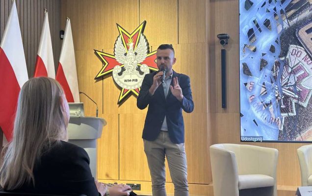 Stojący przed widownią prelegent mówiący do mikrofonu. Za nim na dużym wyświetlaczu fragment prezentacji.