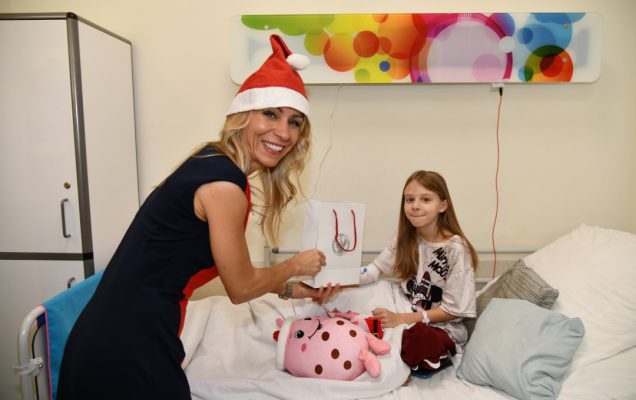 Sala szpitalna. Małgorzata Złotkowska z czapką świętego Mikołaja na głowie wręcza prezent małej pacjentce.