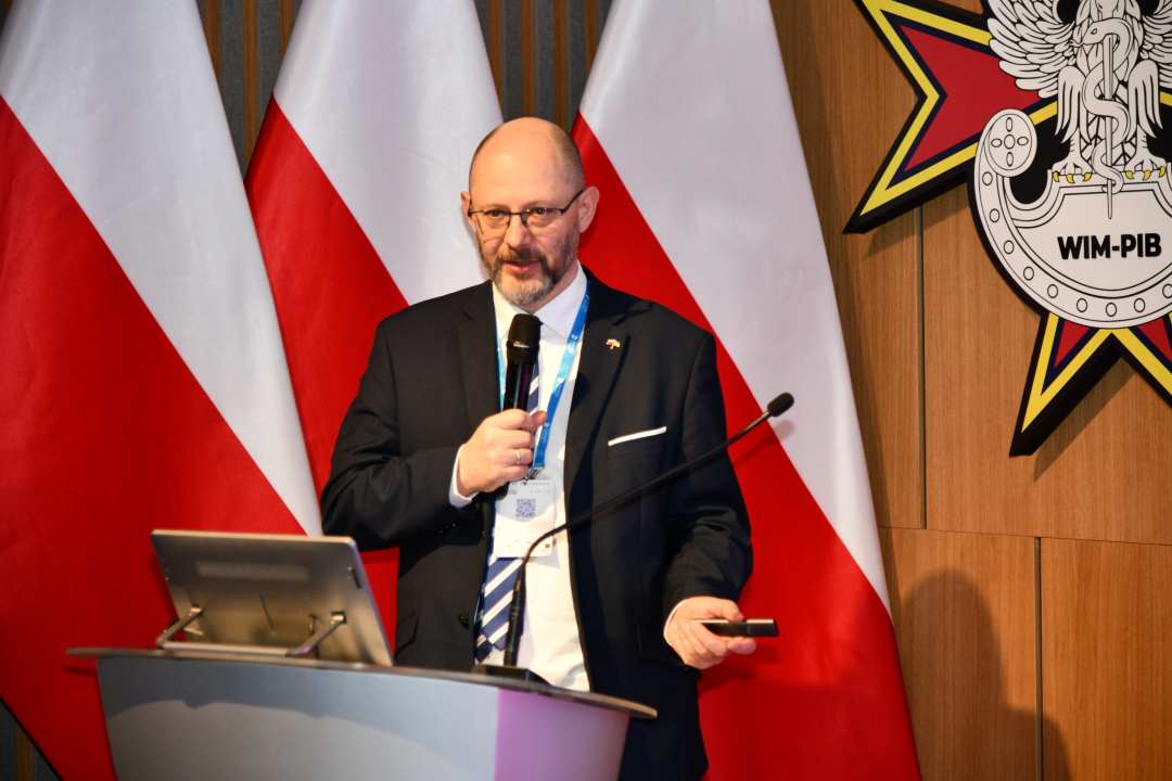 Prelegent przy mównicy, za nim logo WIM i stojące 3 flagi Polski.