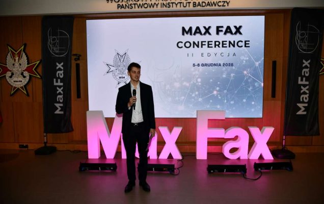 Prelegent podczas konferencji. Za nim podświetlony napis Max Fax i slajd tytułowy konferencji MAxFax.
