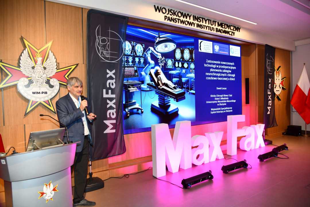 Prelegent przy mównicy. Za nim logo WIM. Obok stoi różowa dekoracja z napisem Max Fax i ekran z wyświetlonym slajdem z konferencji.