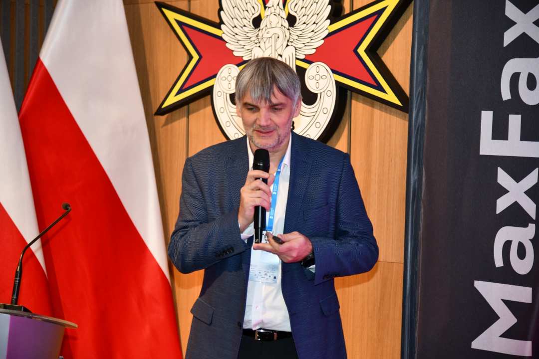 Prelegent z mikrofonem w ręku. Za nim logo WIM i flaga Polski, obok napis Max Fax.