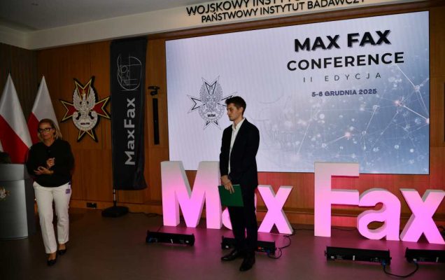 Prelegent podczas konferencji. Za nim podświetlony napis Max Fax i slajd tytułowy konferencji MAxFax. Po prawej stronie doktor Aldona Chloupek.