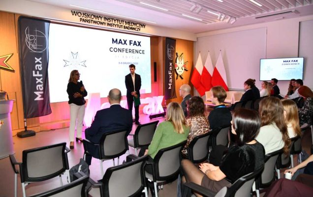 Doktor Aldona Chloupek z Prelegentem podczas konferencji. Za nimi podświetlony napis Max Fax i slajd tytułowy konferencji MAxFax. Przed nimi siedzący słuchacze konferencji.