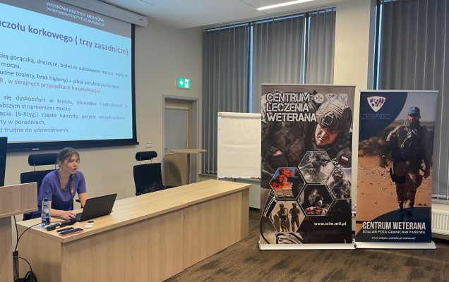 Ppłk dr n. med. Agnieszka Grabińska prowadzi wykład. Za jej plecami plansza wyświetlanej prezentacji. Z boku Stojak z plakatem Centrum leczenie weterana, na plakacie kolaż zdjęć z akcji wojskowych.