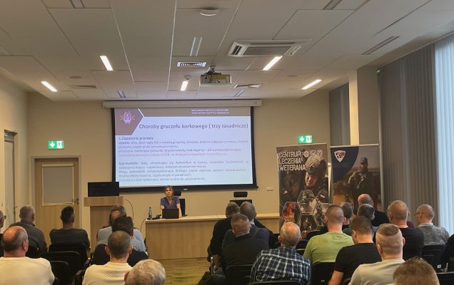 Ujęcie z tyłu na siedzących słuchaczy podczas wykładu. Ppłk dr n. med. Agnieszka Grabińska prowadzi wykład. Za jej plecami plansza wyświetlanej prezentacji. Z boku Stojak z plakatem Centrum leczenie weterana, na plakacie kolaż zdjęć z akcji wojskowych.
