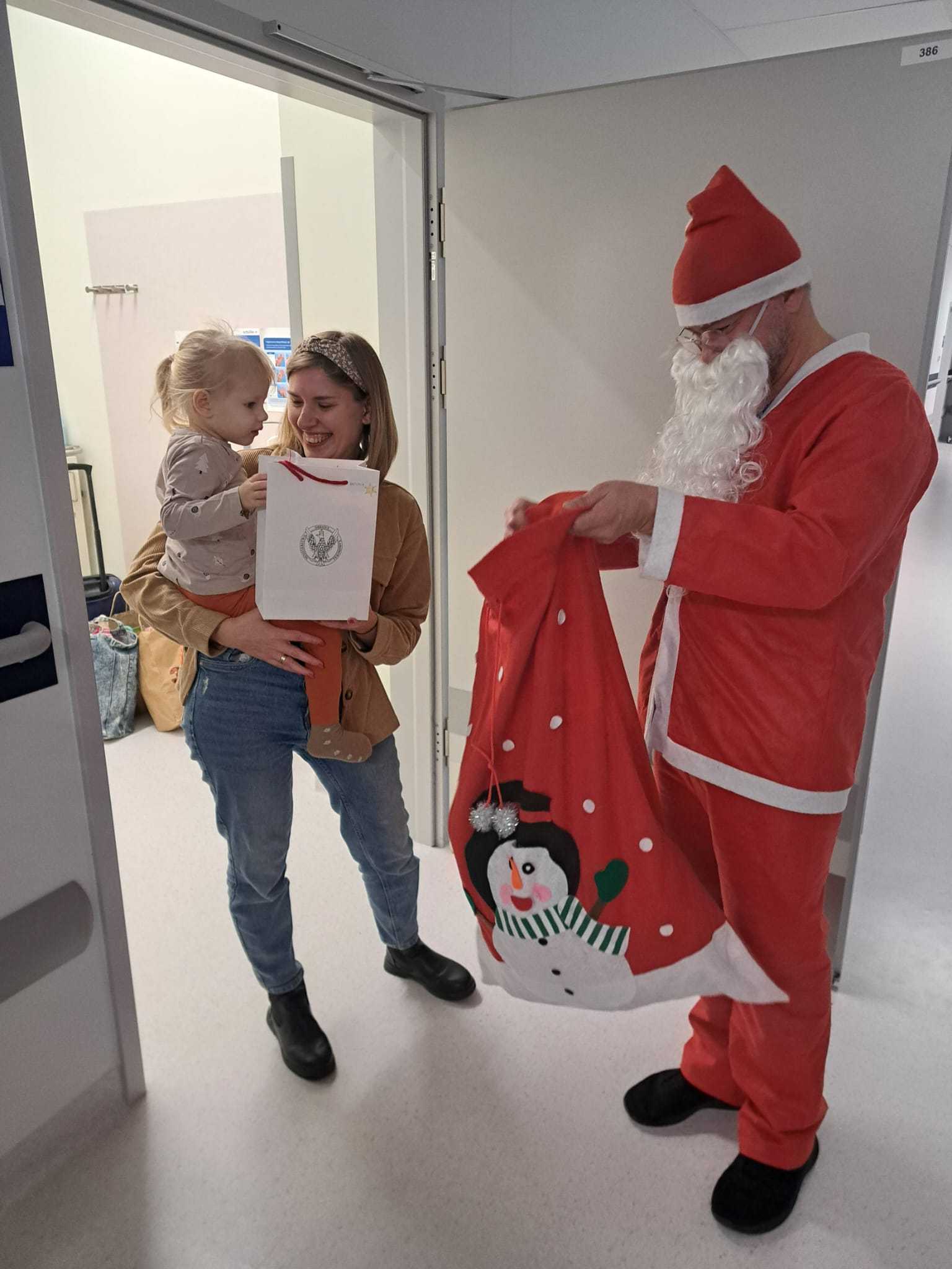 W drzwiach sali stoi uśmiechnięta mama z mała dziewczynką na rękach. Na przeciwko nich stoi Święty Mikołaj i wyjmuje prezent z worka.