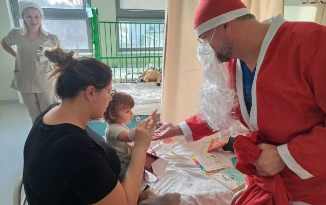 Święty Mikołaj pochyla się nad siedzącą małą dziewczynką i jej mamą przybija piątkę z dziewczynką.
