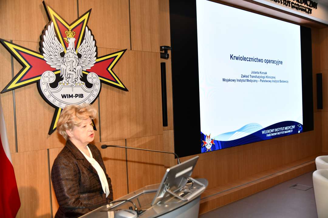 dr hab. n. med. Jolanta Korsak, kierownik Zakładu Transfuzjologii Klinicznej WIM-PIB przy mównicy za jej plecami na ekranie slajd z konferencji.