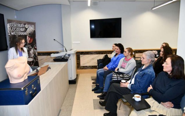 Sala wykładowa. Lekarka prowadząca prelekcję obok niej na katedrze stoi fantom kobiecej klatki piersiowej. Na przeciwko niej siedzące słuchaczki.