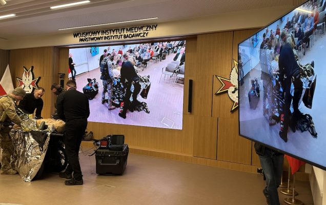 Pokaz ratownictwa medycyny pola walki na auli WIM. Ratownicy przy fantomie poszkodowanego żołnierza. Na ścianach dwa duże monitory wyświetlają relację z pokazu.