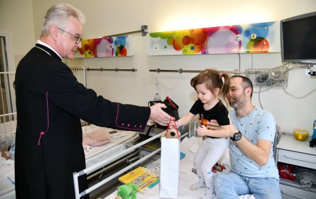 Sala szpitalna. Na łóżku szpitalnym małą dziewczynkę podtrzymuje tata. Dziewczynka odbiera torbę z prezentem od biskupa.