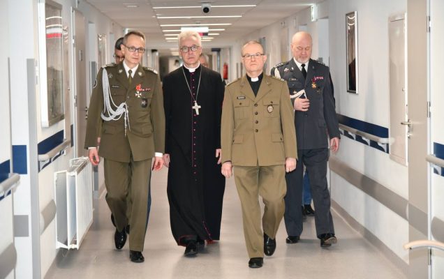 Korytarz szpitalny, biskup z dyrektorem WIM i pracownikami idą do spotkać się z pacjentami.