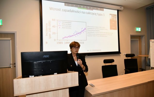 Agnieszka Szypowska z mikrofonem w ręku, podczas wykładu w sali LOMTI. Za nią tablica z prezentacją.