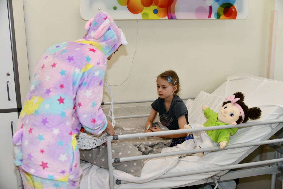 Zajączek wielkanocny w sali szpitalnej wręcza prezent dziewczynce siedzącej na łóżku.
