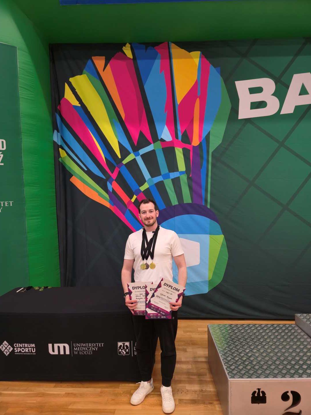 Wojciech Kurzyński na hali sportowej z medalami na szyi, prezentujący dyplomy.