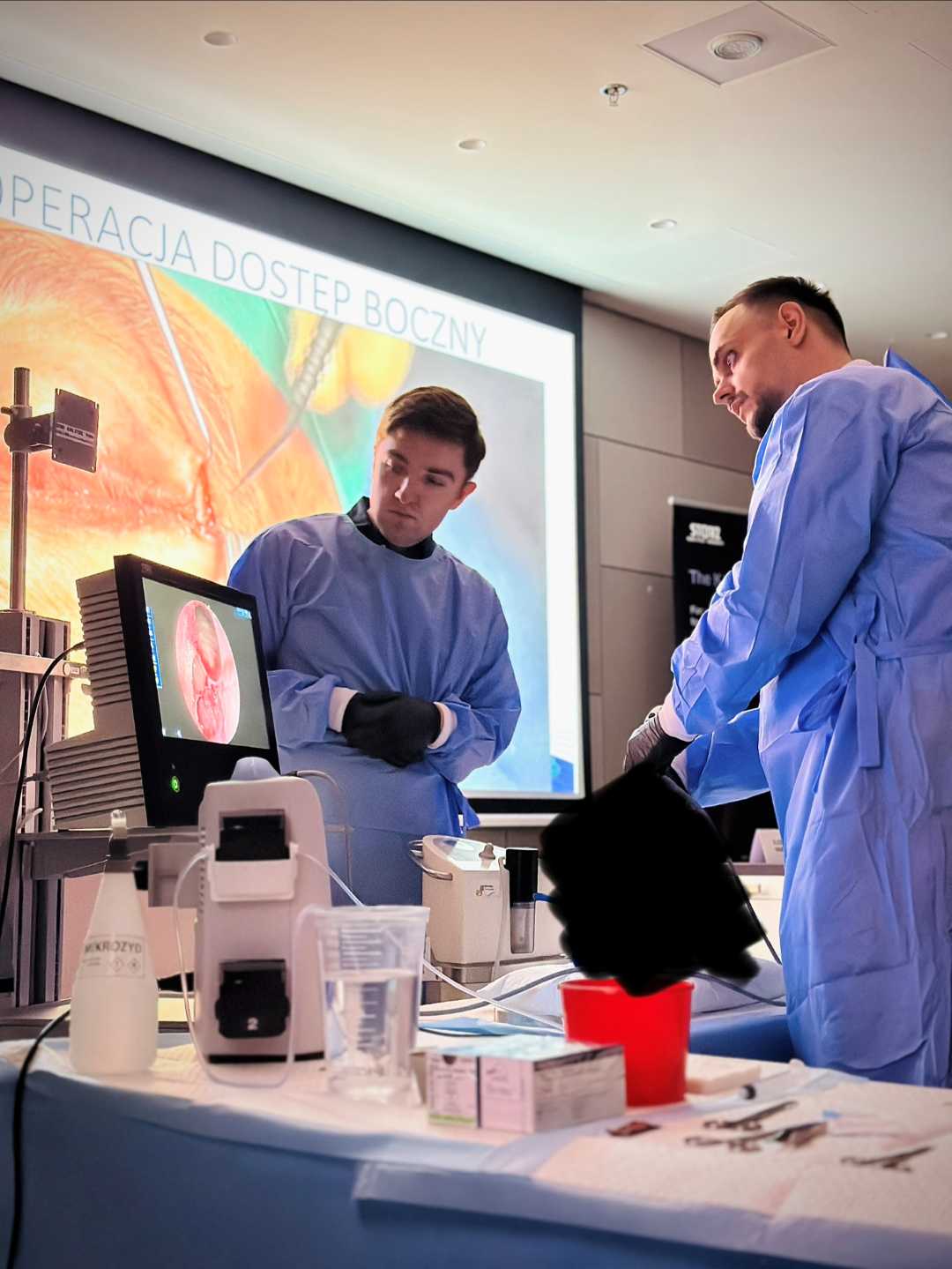 Dwóch młodych mężczyzn w strojach ochronnych stoi przy aparaturze laboratoryjnej. Za ich plecami na ekranie wyświetlona prezentacja.