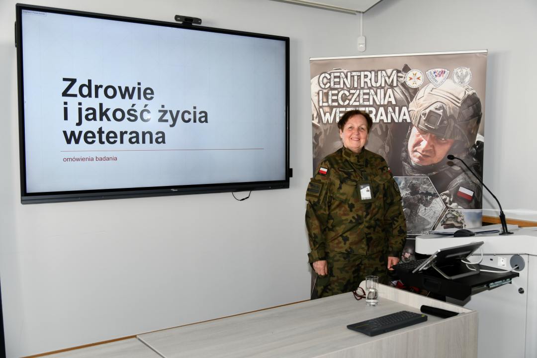 Barbara Betiuk w sali wykładowej. Obok niej na ekranie slajd z prezentacji z napisem Zdrowie i jakość życia weterana. Za jej plecami ścianka reklamowa Centrum leczenia Weterana z żołnierzem podczas ćwiczeń na poligonie.