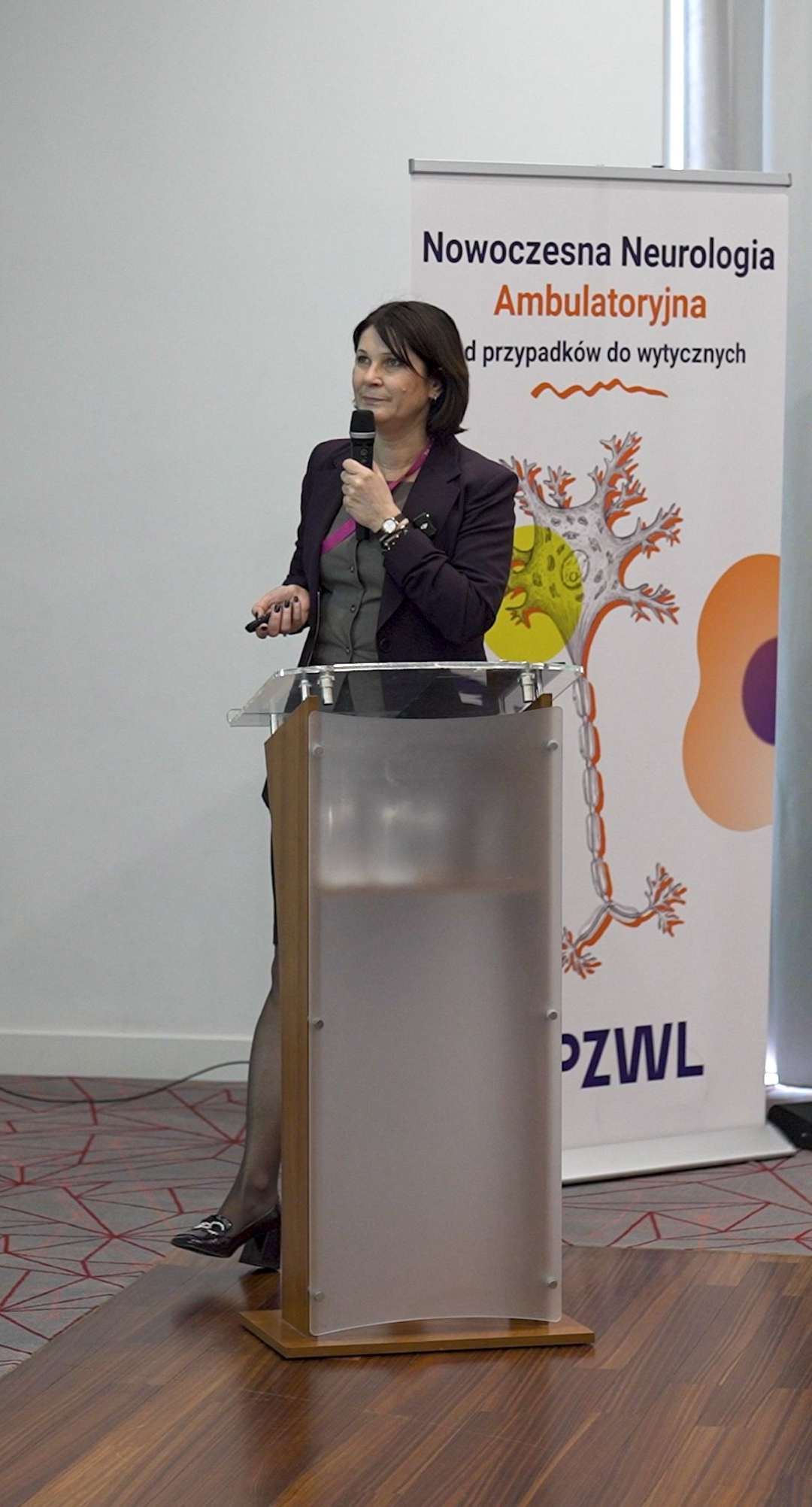 Prelegentka przy mównicy podczas konferencji, za nią ścianka reklamująca konferencję.