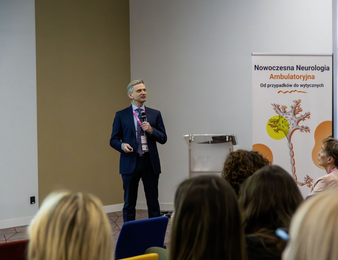 Prelegent przy mównicy podczas konferencji, za nim ścianka reklamująca konferencję.