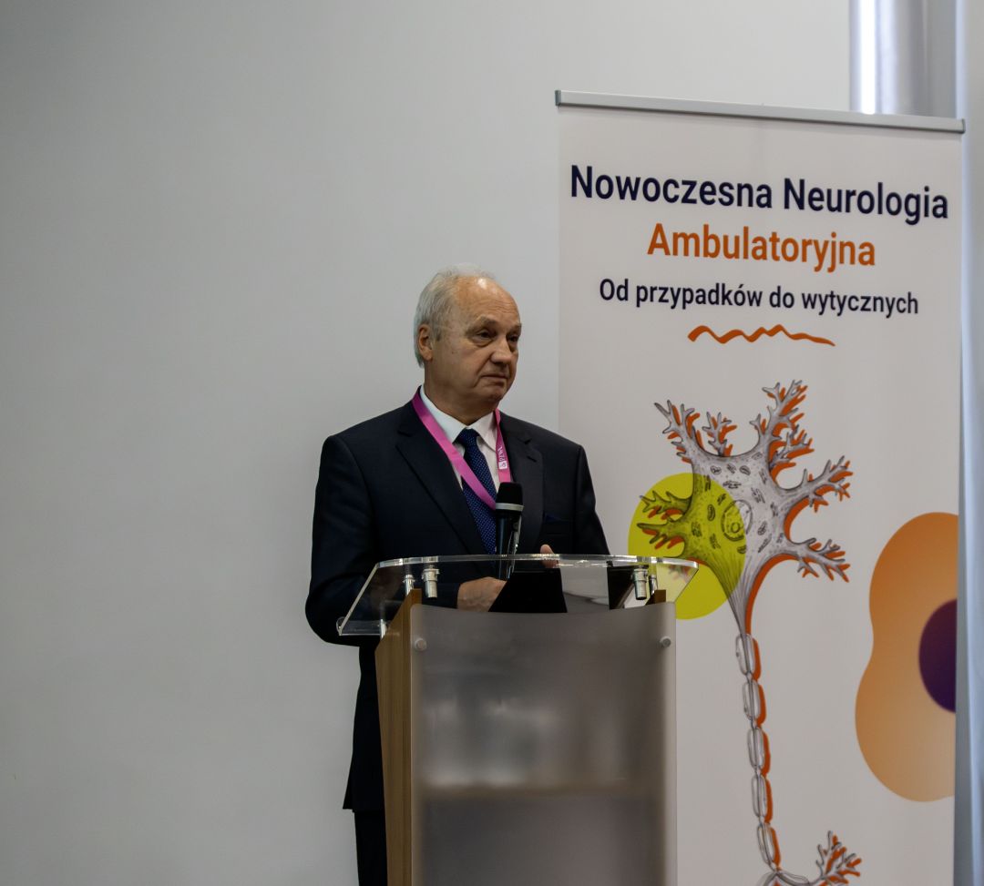 Prelegent przy mównicy podczas konferencji, za nim ścianka reklamująca konferencję.