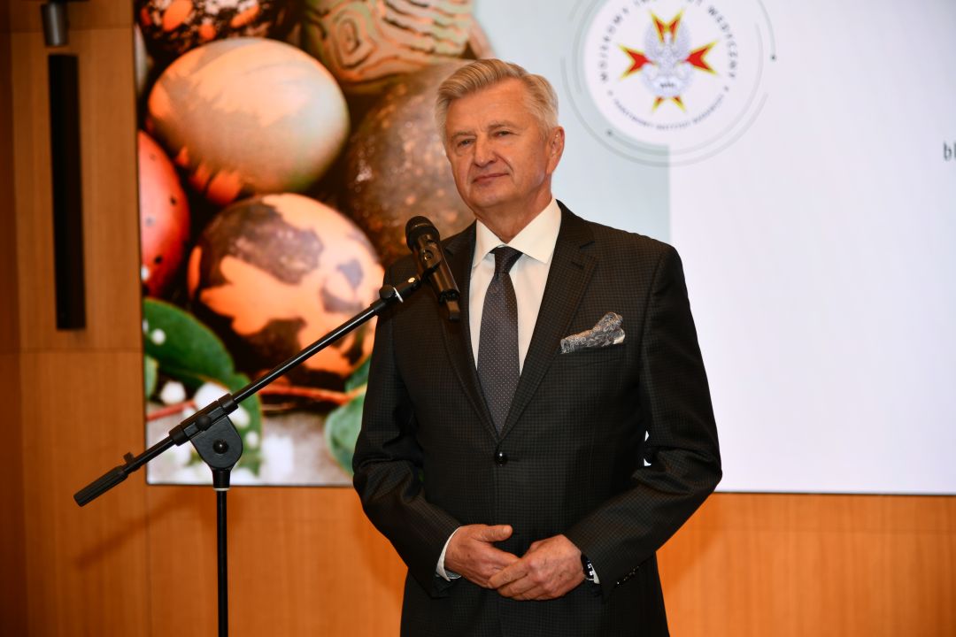 Stanisław Wziątek przy mikrofonie, za nim na ekranie wyświetlone życzenia wielkanocne.