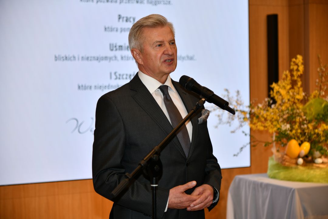 Stanisław Wziątek przy mikrofonie, za nim na ekranie wyświetlone życzenia wielkanocne.