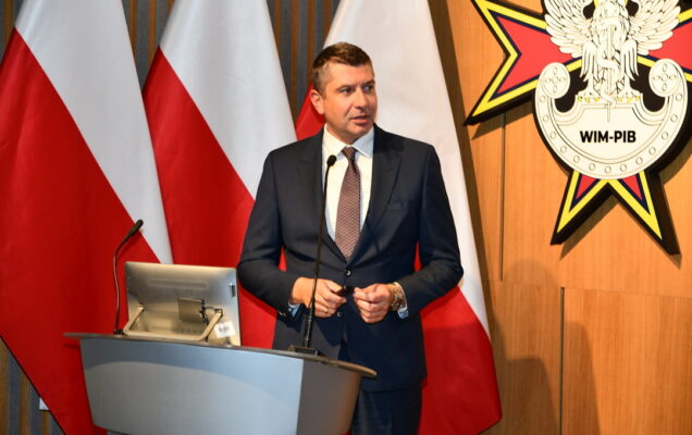 Adrian Szulczyński przy mównicy, za nim trzy flagi Polski oraz logo WIM