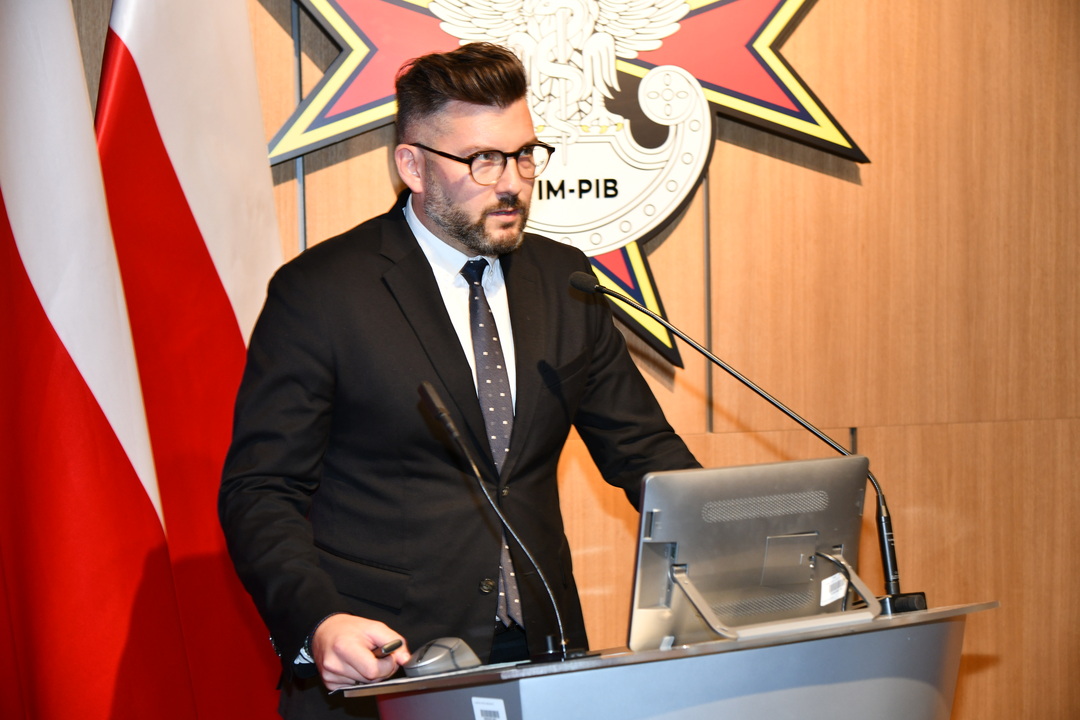 Piotr Chojan przy mównicy, za nim dwie flagi Polski oraz logo WIM